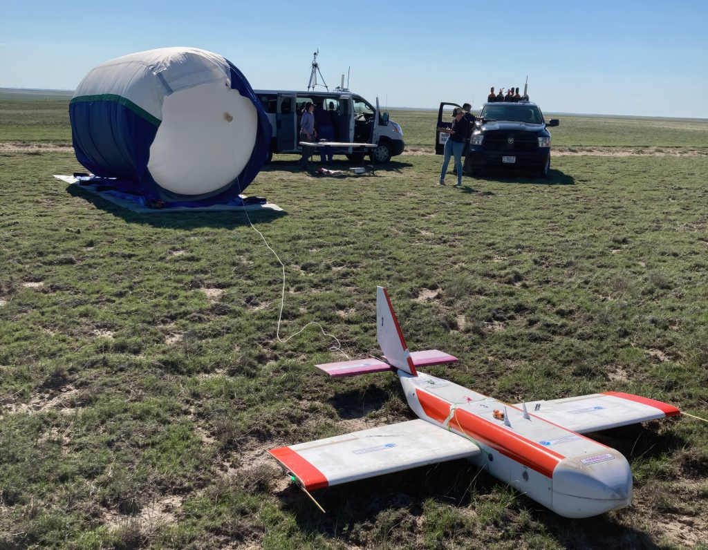 An uncrewed glider sits on the ground in front of a high-altitude balloon and two research vans