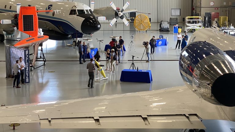 Media members gathered around aircraft and drones inside a hangar