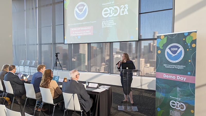 A woman standing in front of a digital screen project and next to a NOAA logo banner addresses an audience.