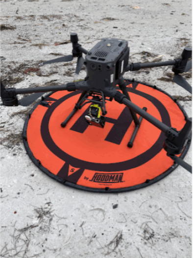 A black drone with an orange base set on a beach.